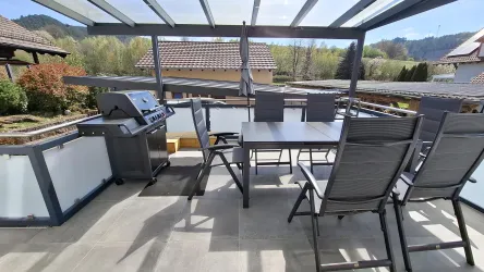 Eine moderne Terrasse mit einem Grill und einem großen Tisch. Die Umgebung ist grün und sonnig, ideal für gesellige Stunden im Freien.