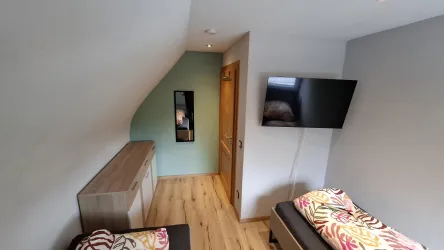 Ein kleines, gemütliches Zimmer mit schrägen Wänden. Es gibt einen Fernseher, ein Bett und einen Schrank aus Holz.