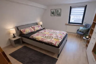 Ein modernes Schlafzimmer mit einem Doppelbett und bunten Bettbezügen. Es gibt einen Stuhl und Bücherregale, die dem Raum eine einladende Atmosphäre verleihen.