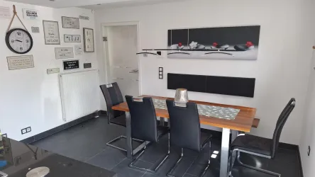 Ein moderner Essbereich mit einem großen Holztisch und schwarzen Stühlen. An der Wand hängen verschiedene Bilder und eine Uhr.