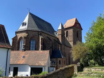 Eine historische Kirche mit roten und grauen Dächern. Umgeben von Bäumen und einem klaren blauen Himmel.