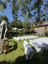 Ein schöner Garten mit weißen Tischen und Bänken unter einem blauen Himmel. Im Hintergrund sind Pavillons und ein Gebäude zu sehen.
