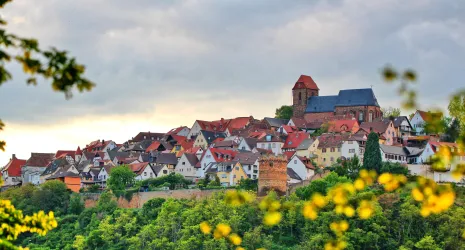 Eine malerische Stadt mit bunten Häusern und einer historischen Kirche auf einem Hügel. Im Vordergrund sind grüne Bäume und eine alte Mauer zu sehen.