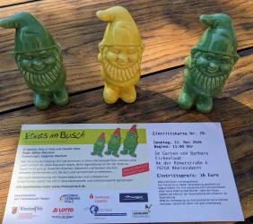 Drei bunte Gartenzwerge stehen auf einem Tisch. Daneben liegt eine Einladung zu einer Veranstaltung.