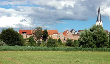 Eine malerische Landschaft mit einem Dorf im Hintergrund und einer Kirche mit spitzem Turm. Der Himmel ist teilweise bewölkt und die Wiesen sind grün.