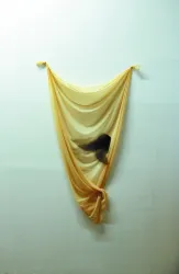 Ein transparentes, gelbes Tuch hängt an der Wand und bildet sanfte Wellen. Dahinter ist eine schemenhafte Gestalt zu erkennen, die eine mysteriöse Atmosphäre schafft.