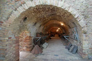 Ein alter, gewölbter Gang mit Ziegelsteinen und sanfter Beleuchtung. An den Wänden sind verschiedene Utensilien und Gegenstände ausgestellt.