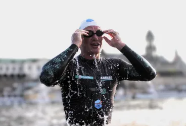 Ein Schwimmer steht am Ufer und zieht seine Schwimmbrille ab. Spritzwasser ist um ihn herum zu sehen.