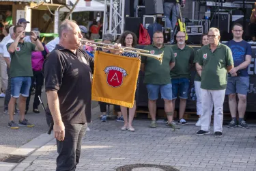 Ein Mann spielt Trompete und hält eine Fahne in der Hand. Im Hintergrund stehen mehrere Personen und schauen zu.