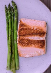 Ein Stück gebratener Lachs liegt auf einem weißen Teller. Daneben befinden sich frische grüne Spargelstangen.