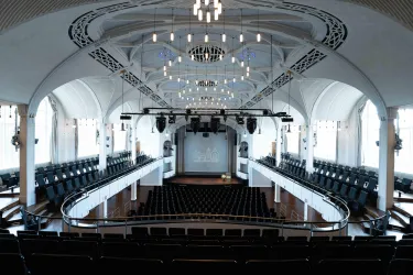 Ein eleganter Veranstaltungsraum mit modernen Sitzplätzen und einer großen Leinwand. Die architektonischen Details verleihen dem Raum eine besondere Atmosphäre.