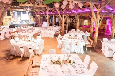 Ein stilvoll dekorierter Veranstaltungsraum mit weißen Tischen und Stühlen. Die Atmosphäre wird durch farbige Beleuchtung und Holzbalken in der Decke unterstrichen.