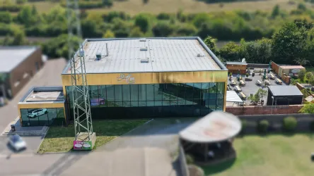 Ein modernes, gelbes Gebäude mit großen Fenstern und einem klaren Himmel. Im Hintergrund sind Sitzbereiche im Freien und grüne Landschaft zu sehen.