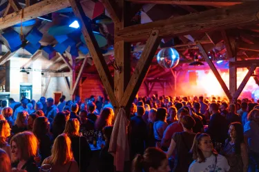 Eine lebhafte Party in einer Holzhütte mit vielen Menschen. Bunte Lichter und eine entspannte Atmosphäre sorgen für gute Stimmung.