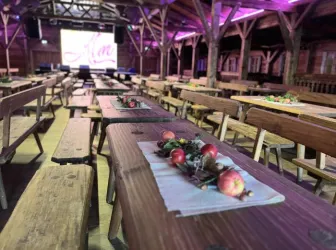 Ein rustikaler Veranstaltungsraum mit langen Holztischen und Bänken. Die Tische sind dekoriert mit frischen Äpfeln und Pflanzen, während im Hintergrund eine Präsentation stattfindet.