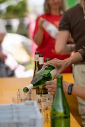 Eine Person gießt grünen Wein in Gläser auf einem Tisch. Im Hintergrund sind weitere Personen und Flaschen sichtbar.