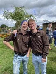 Zwei Männer stehen lächelnd in einem grünen Feld. Beide tragen braune Hemden und Blue Jeans, im Hintergrund sind weitere Menschen und Bäume zu sehen.