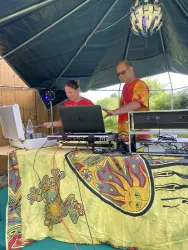 Zwei DJs stehen hinter einem bunten Mischpult unter einem Zelt. Sie sind konzentriert bei der Arbeit, während im Hintergrund eine entspannte Stimmung herrscht.