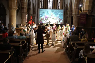 Eine Aufführung in einer Kirche mit vielen Kindern, die in bunten Kostümen stehen. Im Hintergrund ist eine große Leinwand mit einer bunten Szene zu sehen.