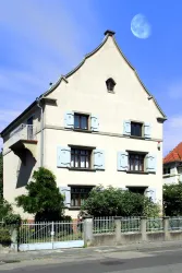 Ein charmantes Haus mit blauen Fensterläden und einem einladenden Garten. Der klare Himmel und der Mond verleihen der Szene eine besondere Atmosphäre.