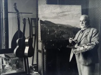 Ein Künstler steht in einem Atelier mit einem Malerpinsel und einer Palette in der Hand. Im Hintergrund sind Gitarren und ein Gemälde mit einer Landschaft zu sehen.