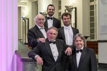 Fünf Männer in eleganter Abendgarderobe stehen lächelnd zusammen. Die Szene wirkt festlich und gehoben.
