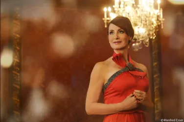 Eine elegante Frau in einem roten Abendkleid steht vor einem glitzernden Kronleuchter. Der Hintergrund ist warm und einladend, was eine festliche Atmosphäre schafft.