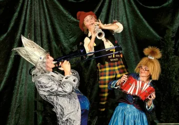 Drei clownartige Musiker in bunten Kostümen spielen verschiedene Instrumente. Die Atmosphäre ist fröhlich und heiter.