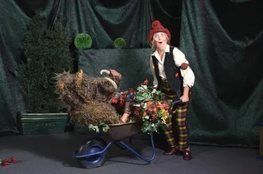 Ein als tapferes Schneiderlein verkleideter Mann mit einem roten Hut schiebt eine Schubkarre, in der ein Wildschwein mit Pflanzen und Blumen sitzt. Der Hintergrund ist grün und dekorativ mit kleinen Bäumen.