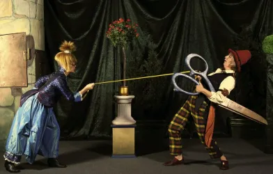 Eine märchenhafte Szene mit zwei Figuren, die an einem langen Band ziehen. Die eine Figur trägt ein blaues Kleid, während die andere einen Hut und eine große Schere hält.