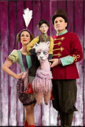 Eine bunte Kunstszene mit zwei Darstellern in Zirkus Kostümen. Sie stehen vor einem rosa Hintergrund und der Mittelpunkt ist ein verkleidetes Lama.