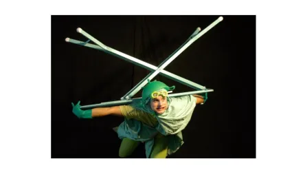 Ein kreatives Kostüm mit grünen Farben und Accessoires. Die Person posiert dynamisch mit weißen Stäben vor einem schwarzen Hintergrund.