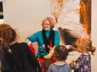 Eine Frau interagiert mit Kindern, während sie Puppen oder Spielzeuge zeigt. Die Atmosphäre ist freundlich und einladend.