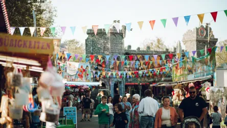 Ein belebter Festplatz mit bunten Fahnen und Ständen. Menschen bewegen sich zwischen den Attraktionen und genießen die festliche Atmosphäre.