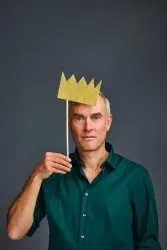 Ein Mann steht vor einem grauen Hintergrund und hält einen goldenen Kronen-Stick in der Hand. Er trägt ein grünes Hemd und hat einen neutralen Gesichtsausdruck.