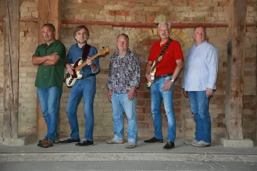 Eine Band mit fünf Musikern steht vor einer gemauerten Wand. Sie halten Instrumente und tragen lässige Kleidung.