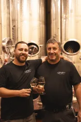 Zwei Männer stehen in einer Weinproduktionsanlage und halten Weingläser in der Hand. Sie lächeln und wirken zufrieden.