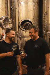 Zwei Männer stoßen mit Weingläsern an, während sie in einem Weinkeller stehen. Im Hintergrund sind große Edelstahltanks zu sehen.