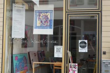 Ein Kunstgeschäft mit bunten Bildern im Schaufenster. Die Fenster sind klar, und mehrere Plakate und Informationen sind sichtbar.