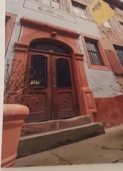 Eine alte, rustikale Holztür mit schmiedeeisernen Gitterelementen. Der Eingang ist von bunten Wänden und einem blauen Himmel umgeben.