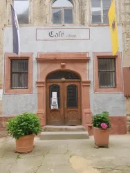 Ein Café mit einer einladenden Fassade aus Stein und Holz. Es gibt Pflanzen in Töpfen vor dem Eingang und Fahnen an den Seiten.