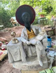 Eine Statue eines sitzenden Mannes in eleganter Kleidung mit einem großen Hut. Er sitzt an einem Tisch mit einer Kugel und ist von einer grünen Umgebung umgeben.