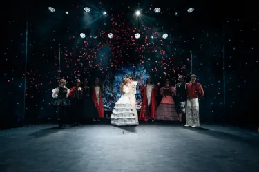 Eine Theateraufführung mit mehreren Schauspielern in historischen Kostümen. Roseblätter fallen vom Himmel, während eine Hauptfigur im weißen Kleid im Mittelpunkt steht.