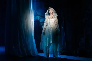 Eine Frau in einem weißen Kleid steht im Scheinwerferlicht. Der Hintergrund ist dunkel und mystisch, mit leichter Beleuchtung.