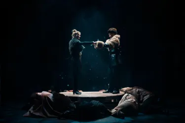 Eine dramatische Bühnenaufführung mit zwei Schauspielern, die sich über eine Bühne greifen. Im Hintergrund liegen mehrere Personen in Kostümen.