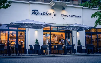 Ein einladendes Café mit dem Namen "Reuther's Backstube". Vor dem Gebäude stehen Tische und Stühle unter großen Sonnenschirmen.
