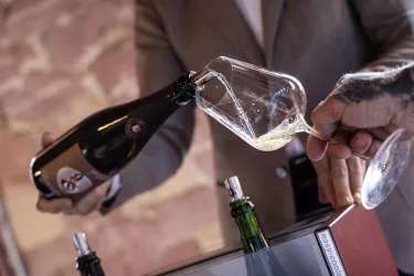 Eine Hand gießt Sekt aus einer Flasche in ein Glas. Im Hintergrund sind weitere Flaschen und ein stilvolles Ambiente zu sehen.