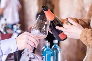 Eine Hand gießt Sekt in ein Glas. Im Hintergrund sind Flaschen und Gläser sichtbar.
