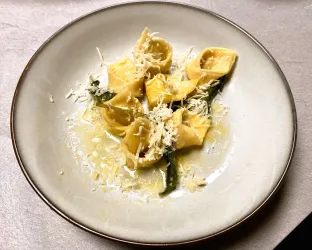 Ein Teller mit frischen Tortellini, garniert mit Käse und grünen Blättern. Die Pasta schwimmt in einer leichten Soße.