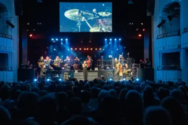 Ein Live-Konzert mit einer Band auf der Bühne. Die Zuschauer im Vordergrund genießen die Musik unter bunten Lichtern.